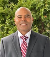 Mass. State Representative Kip Diggs - a Black man in a gray suit, red-and-white striped tie in front of green bushes