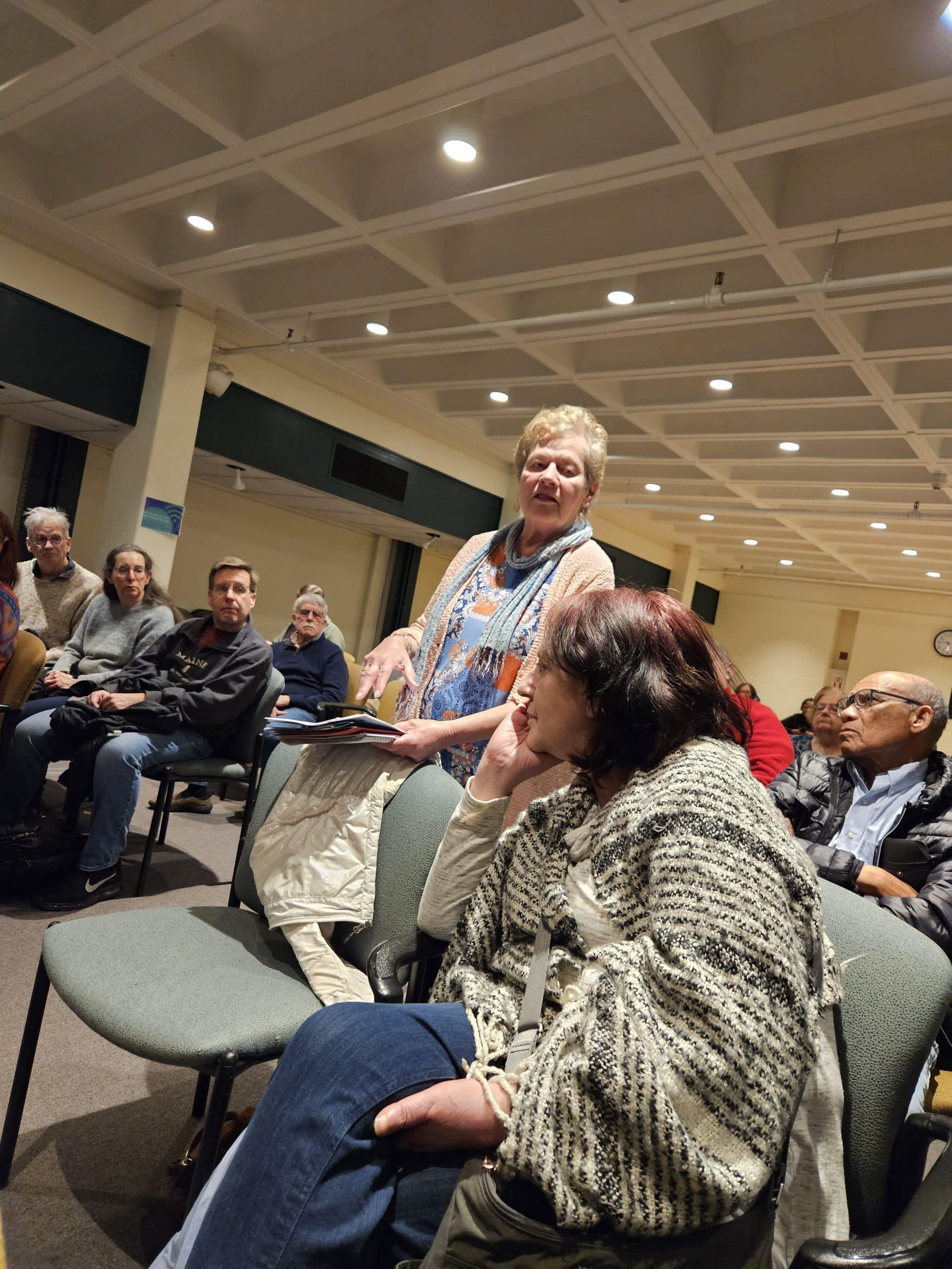 A woman in blue top with orange decorations and beige sweater addresses group